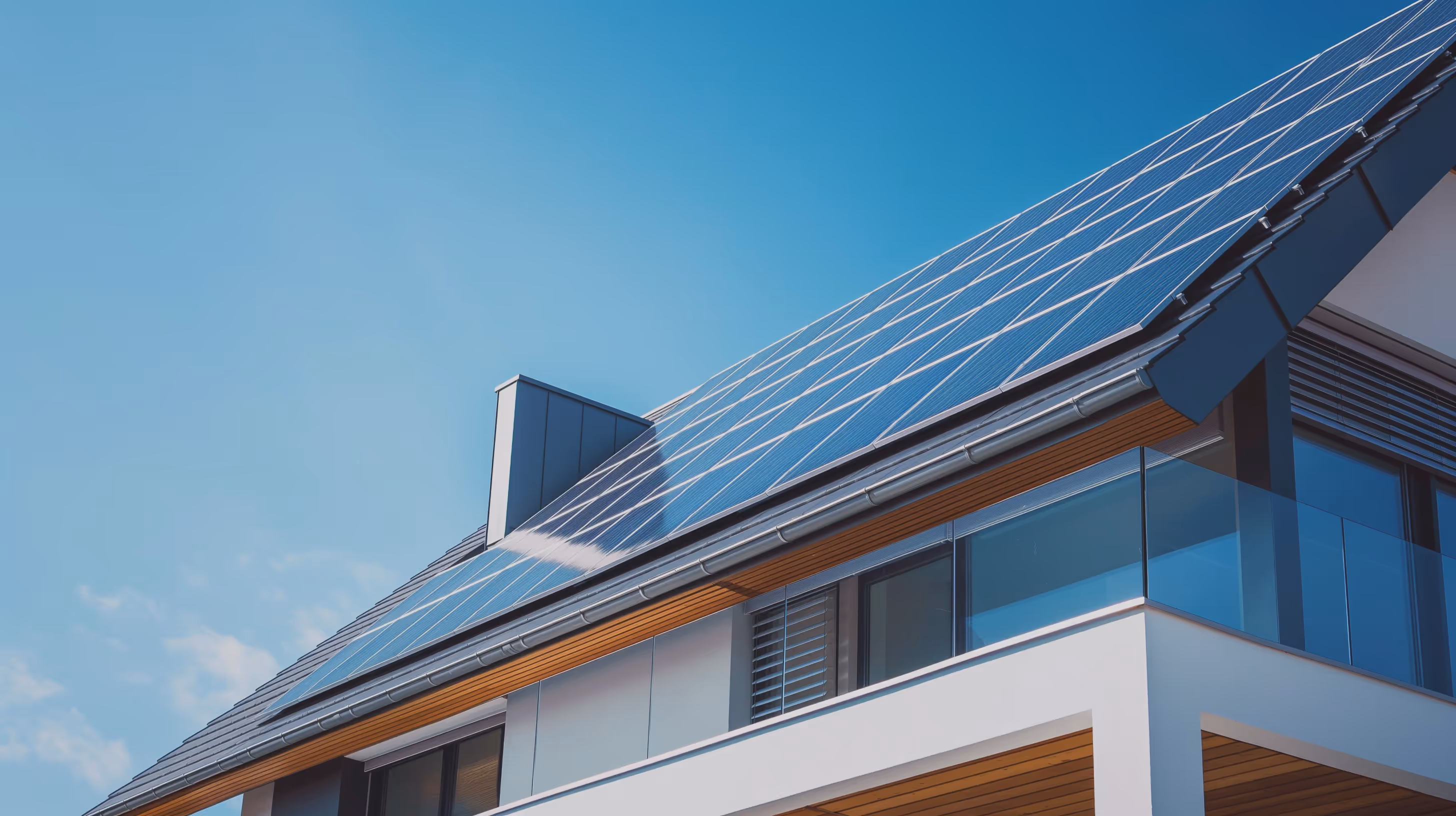 Современный дом с солнечными панелями на крыше и стеклянным балконом на фоне голубого неба.