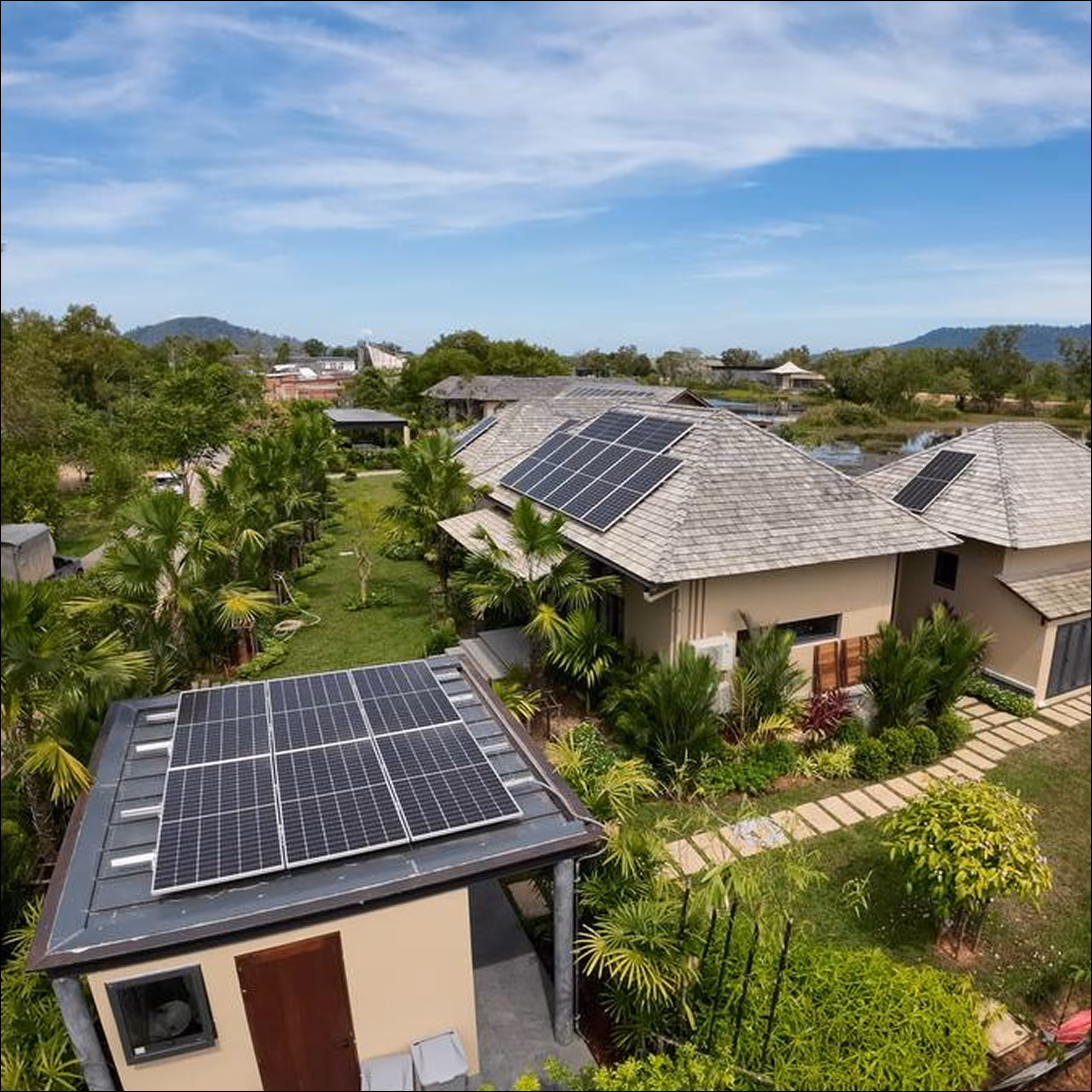 Жилой дом с солнечными панелями на крыше, окружённый зелёными деревьями и растениями под голубым небом.