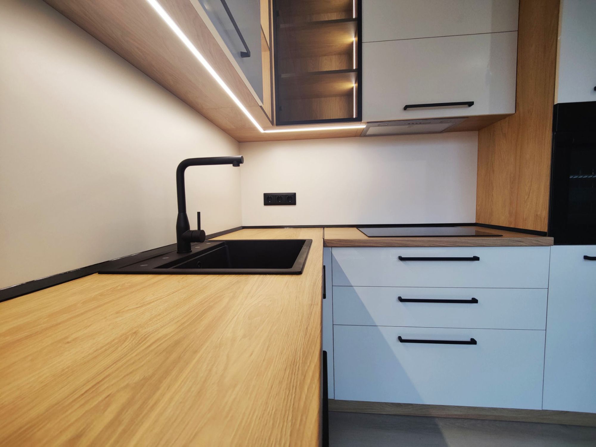 Близък кадър на мивка и плот с черен смесител, LED лента под горните шкафове; плот EGGER H3730 ST10, лице Kronospan 0564 HG/BS, корпус EGGER H3730 ST10, гръб 0564 PE.