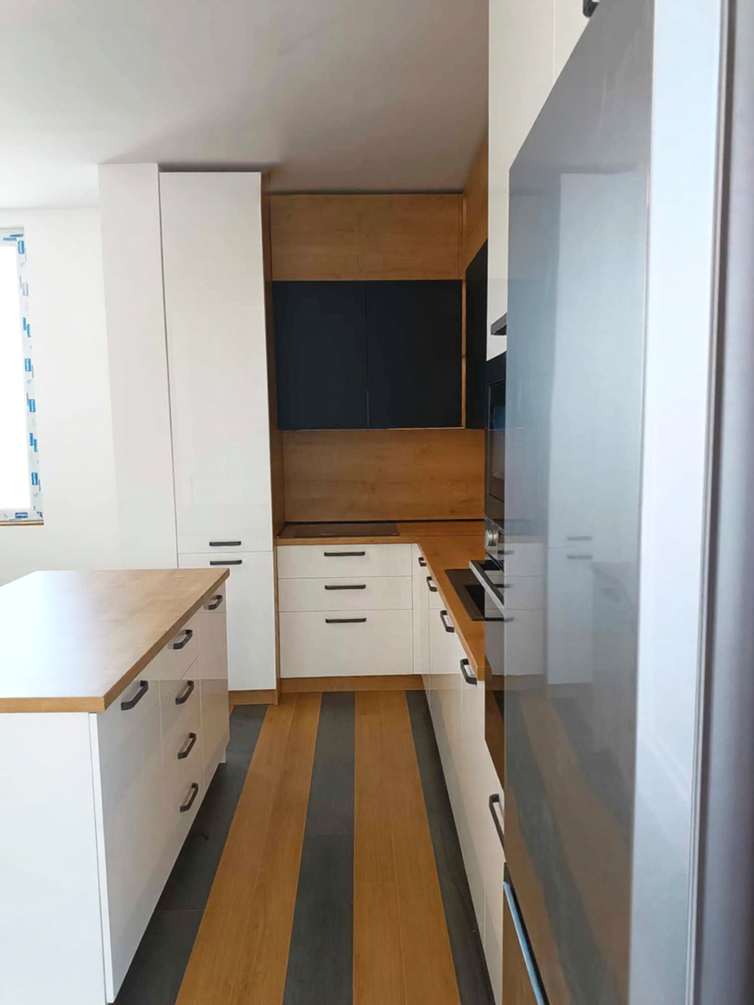 Широк кадър с остров и колона с фурна/микровълнова; гланцово бяло и матово черно, отворени рафтове в острова.