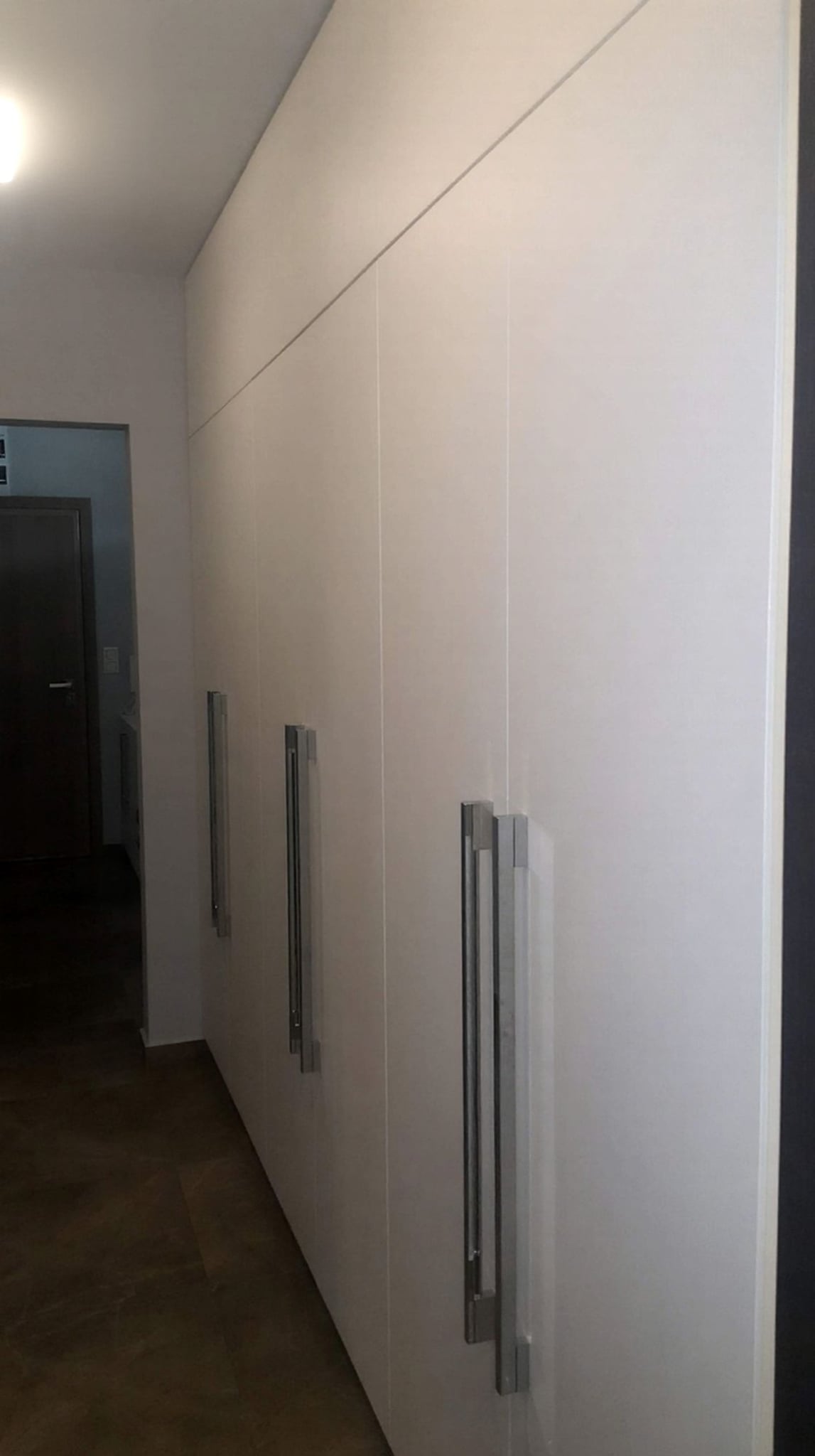 Високи гардеробни шкафове в коридор с гладки бели лица и акцент от тъмен дървесен декор, проектирани от пода до тавана.