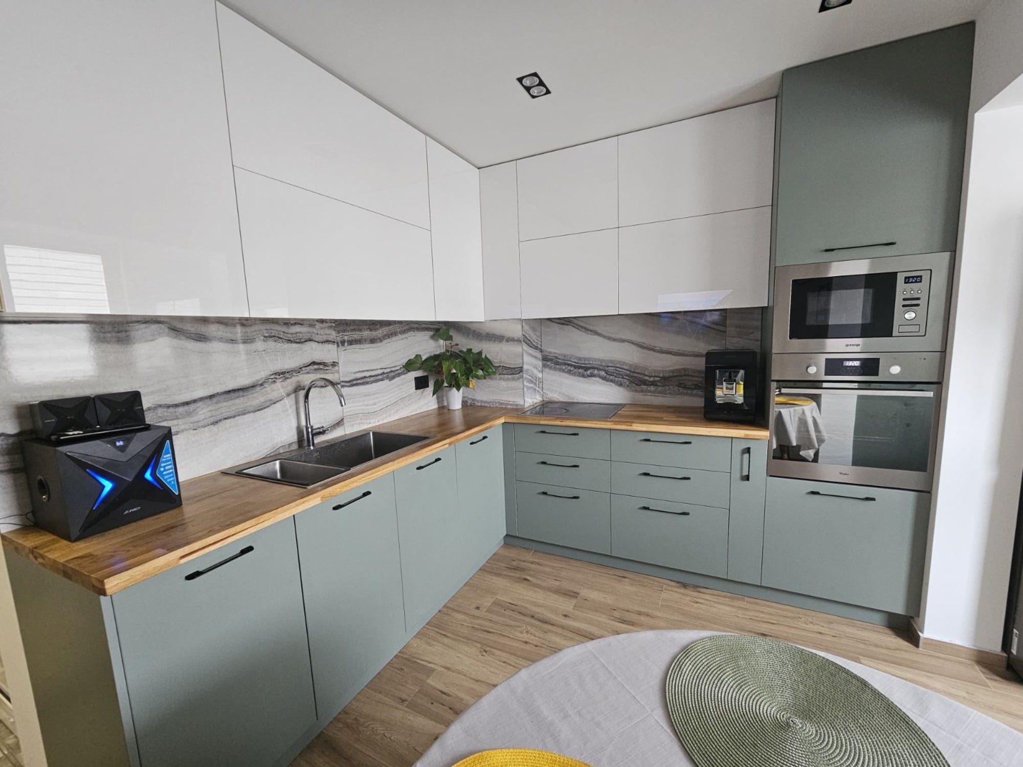 Общ изглед на ъглова кухня с контраст между гланцово бяло и приглушено зелено, масивен плот и естествен под.