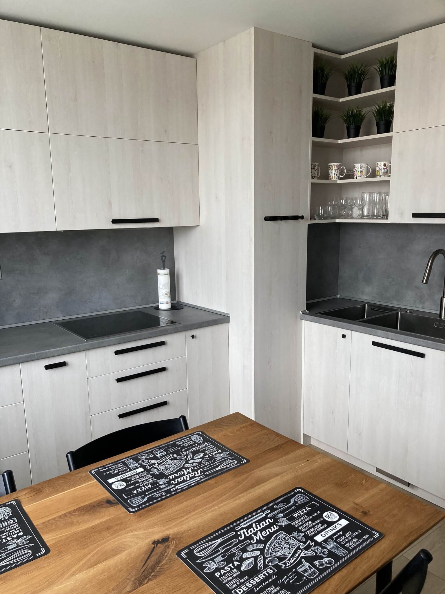 Ъглова модерна кухня по поръчка със светли гланцови шкафове, плот светъл дъб, черни дръжки и LED осветление.