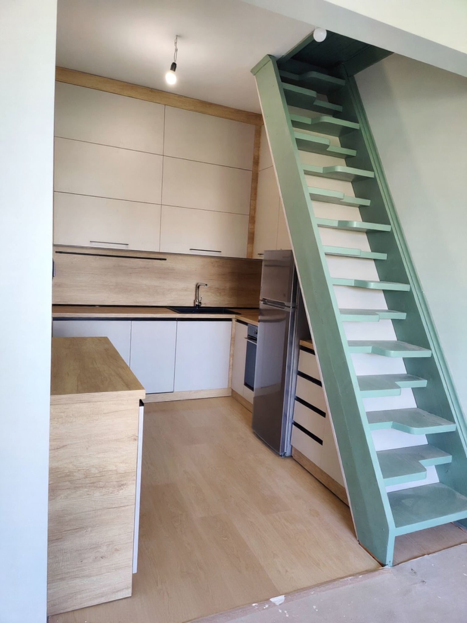 Общ изглед към кухнята под стълбата с L-образен работен плот и LED осветление по стенния панел.