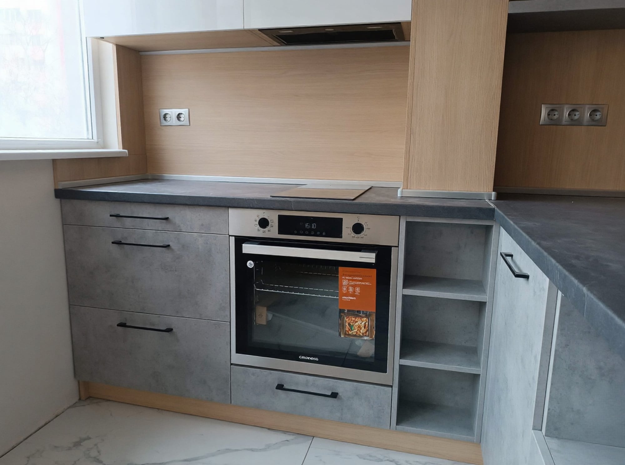 Работна зона с фурна и котлони под прозорец, гръб Дъб Виченца H3157 ST12 и долни шкафове в светъл бетон.