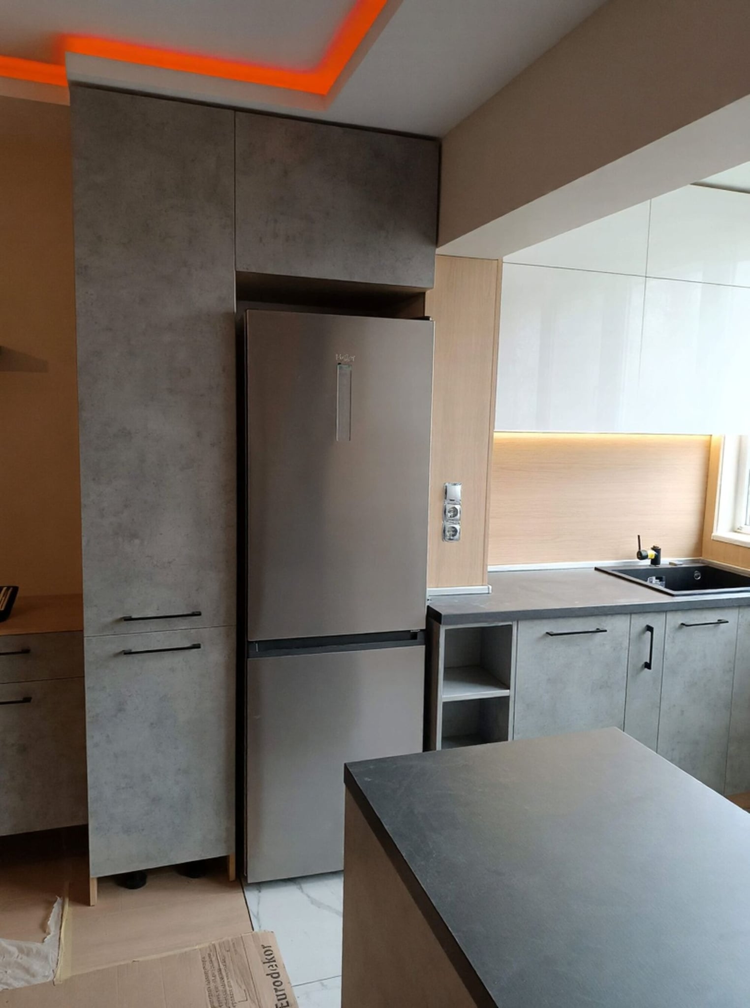 Висок шкаф за хладилник с интегрирани чекмеджета и черни дръжки, контраст между бетонна текстура и дървесен корпус.