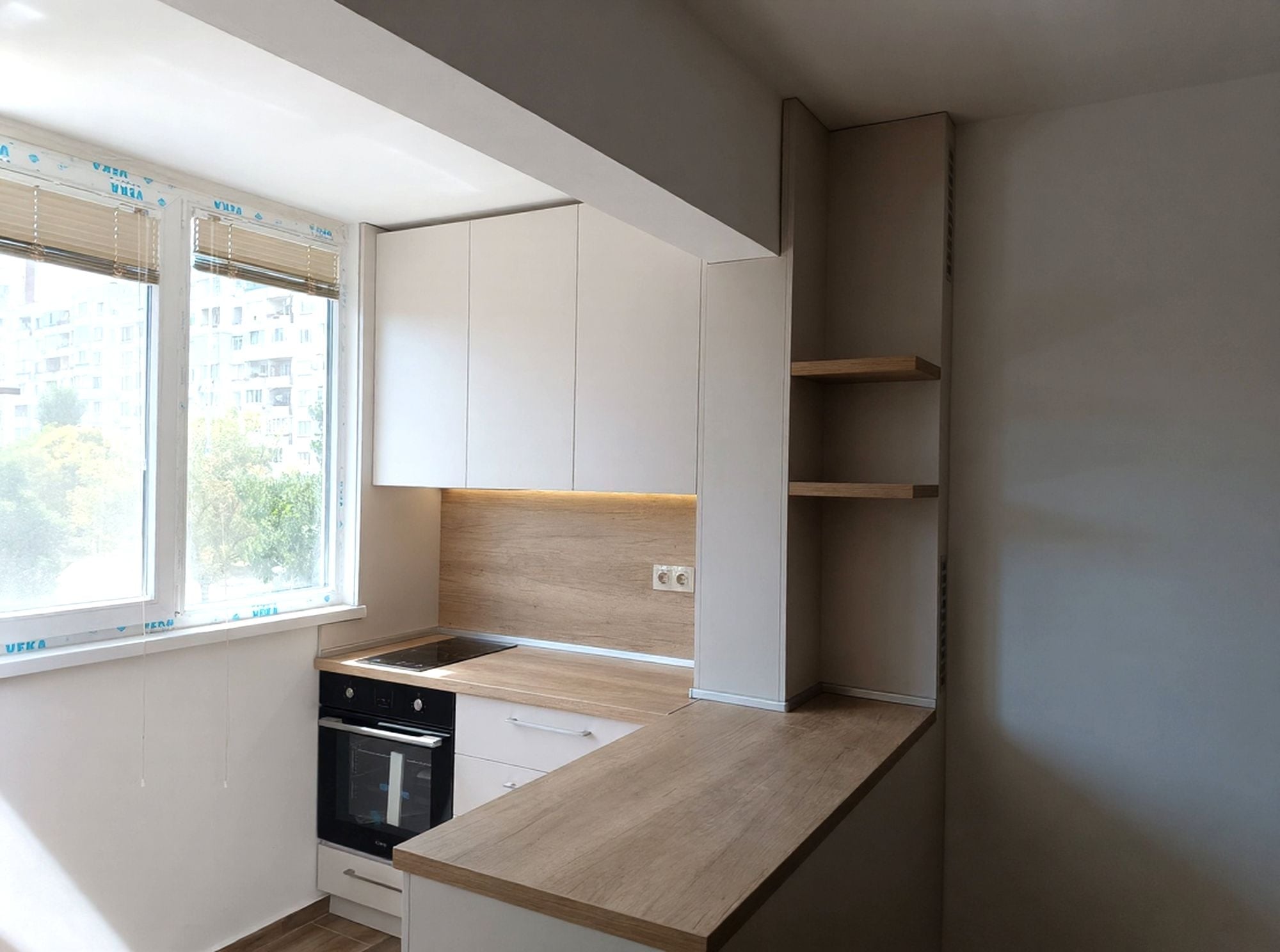 Кухня по поръчка в декор Сива Ангора EGGER с плот и гръб Дъб Небраска натур. Компактно ъглово решение с бар плот към дневната зона, оптимизирано за малки пространства и максимална функционалност.