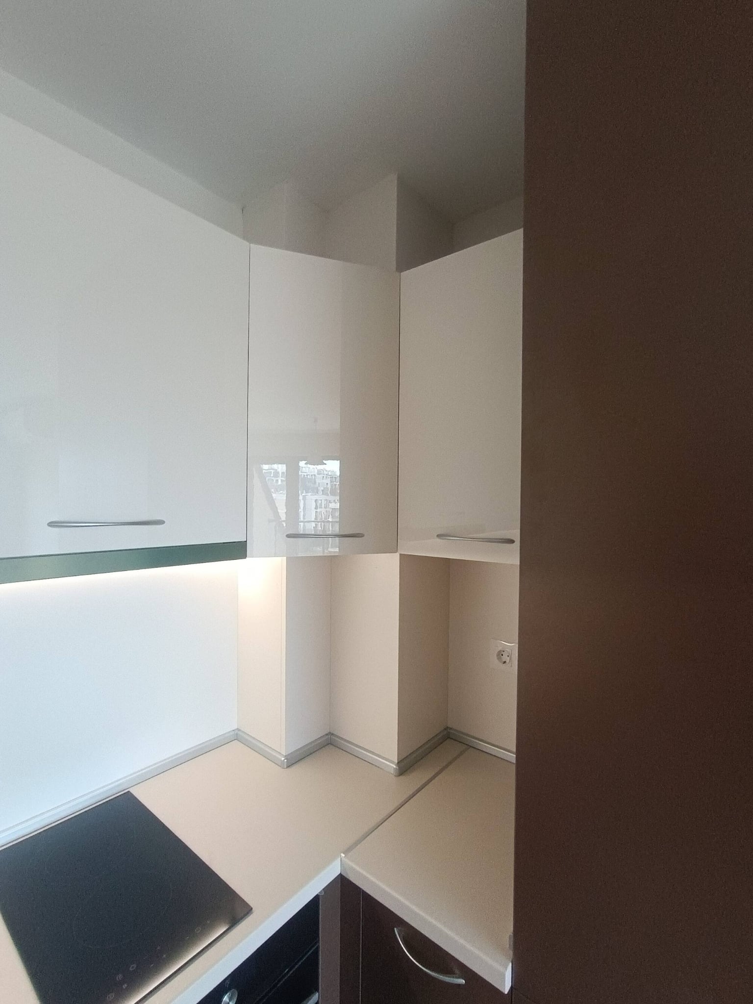 Ъглово решение с непрекъснат бял гръб и LED осветление под горните шкафове, акцент върху гланцовите бадемови врати.