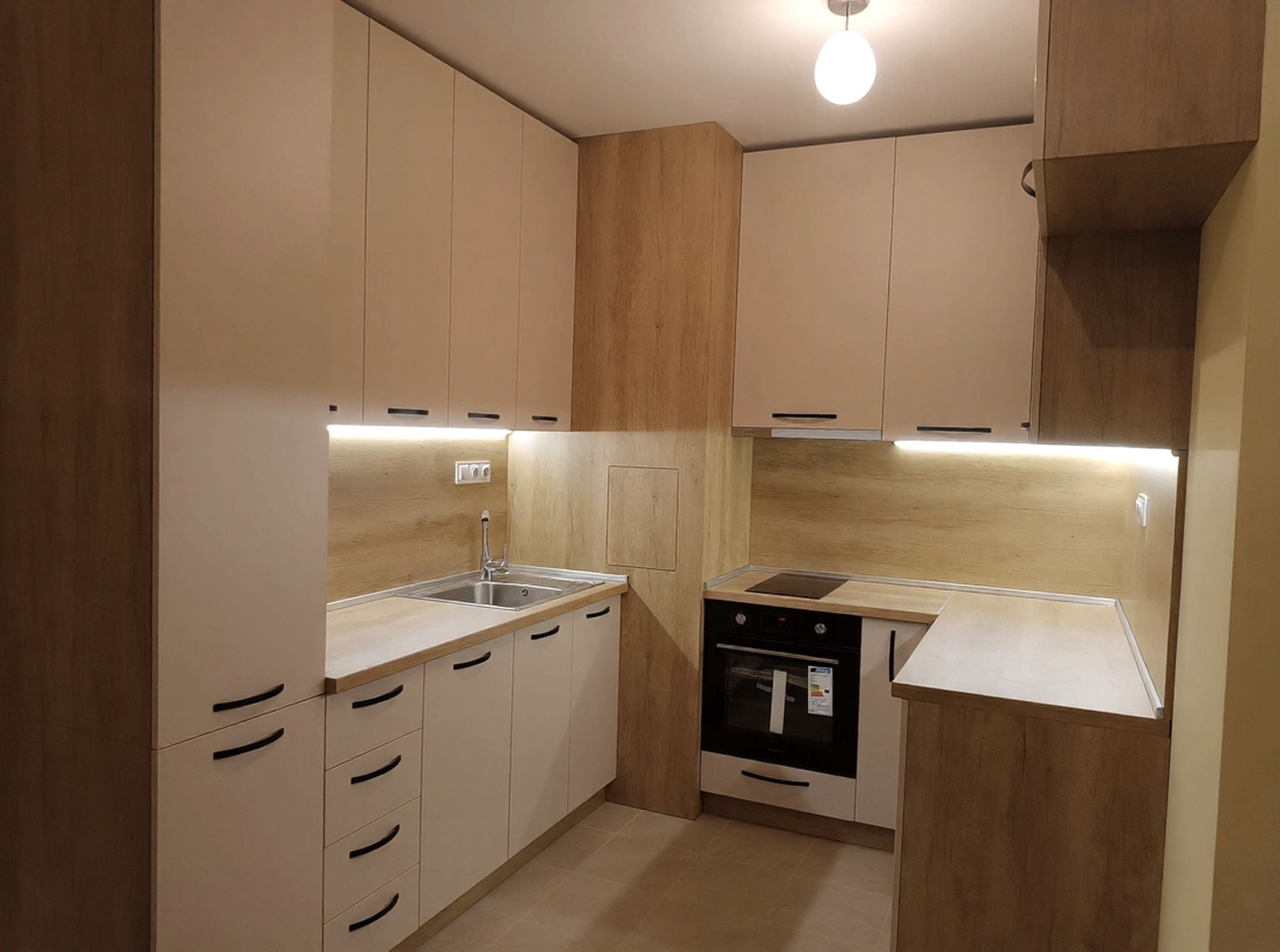 Ъглова кухня по поръчка в топъл дървесен декор H3331 ST10 Дъб Небраска Натур с лица в пясъчно бежово. Компактно решение с много място за съхранение, LED осветление и практичен ревизионен отвор.