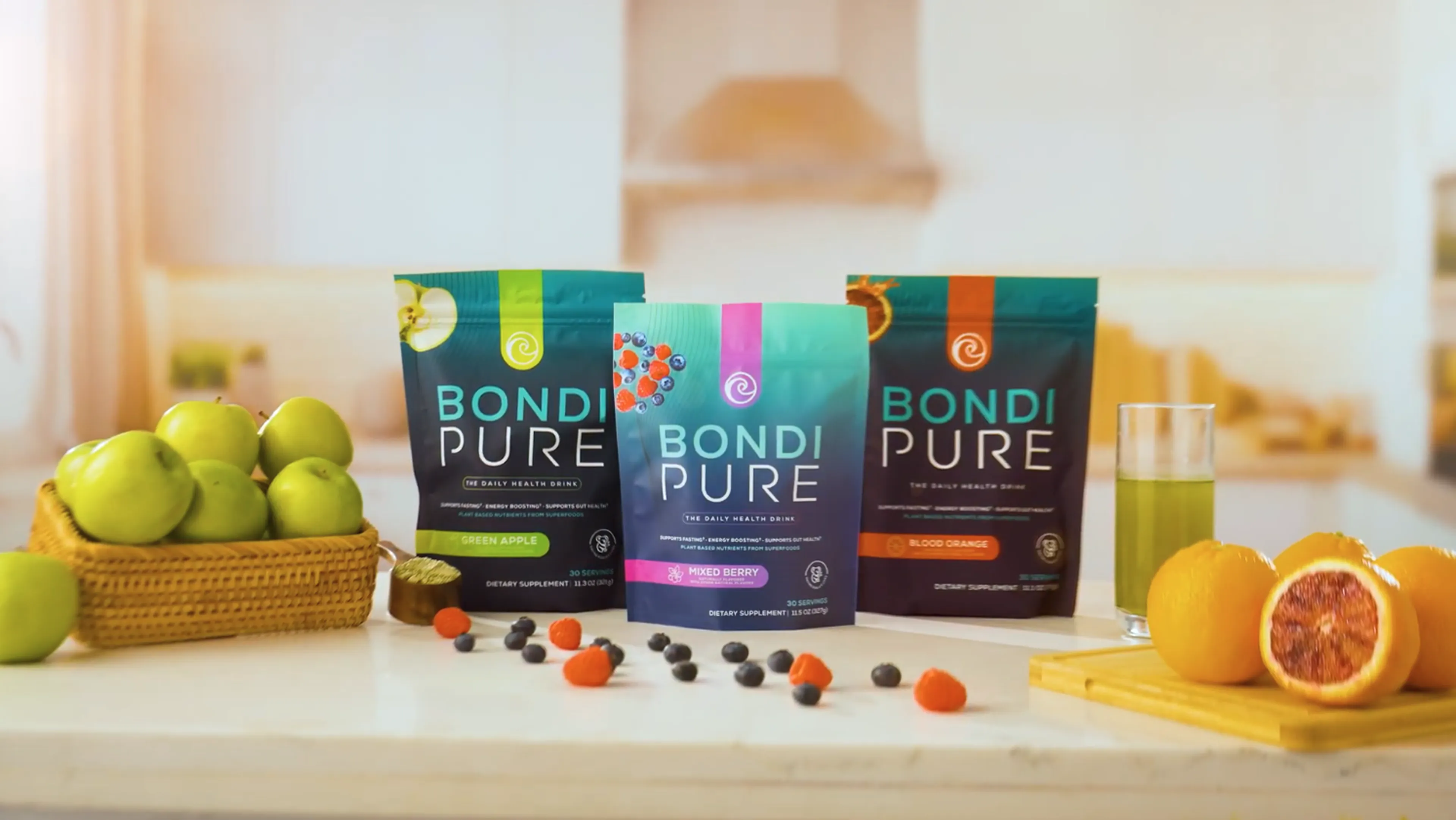 Three Bondi Pure dietary supplement packages—Green Apple, Mixed Berry, and Blood Orange—arranged on a kitchen counter with fresh green apples, berries, blood oranges, and a glass of green juice.