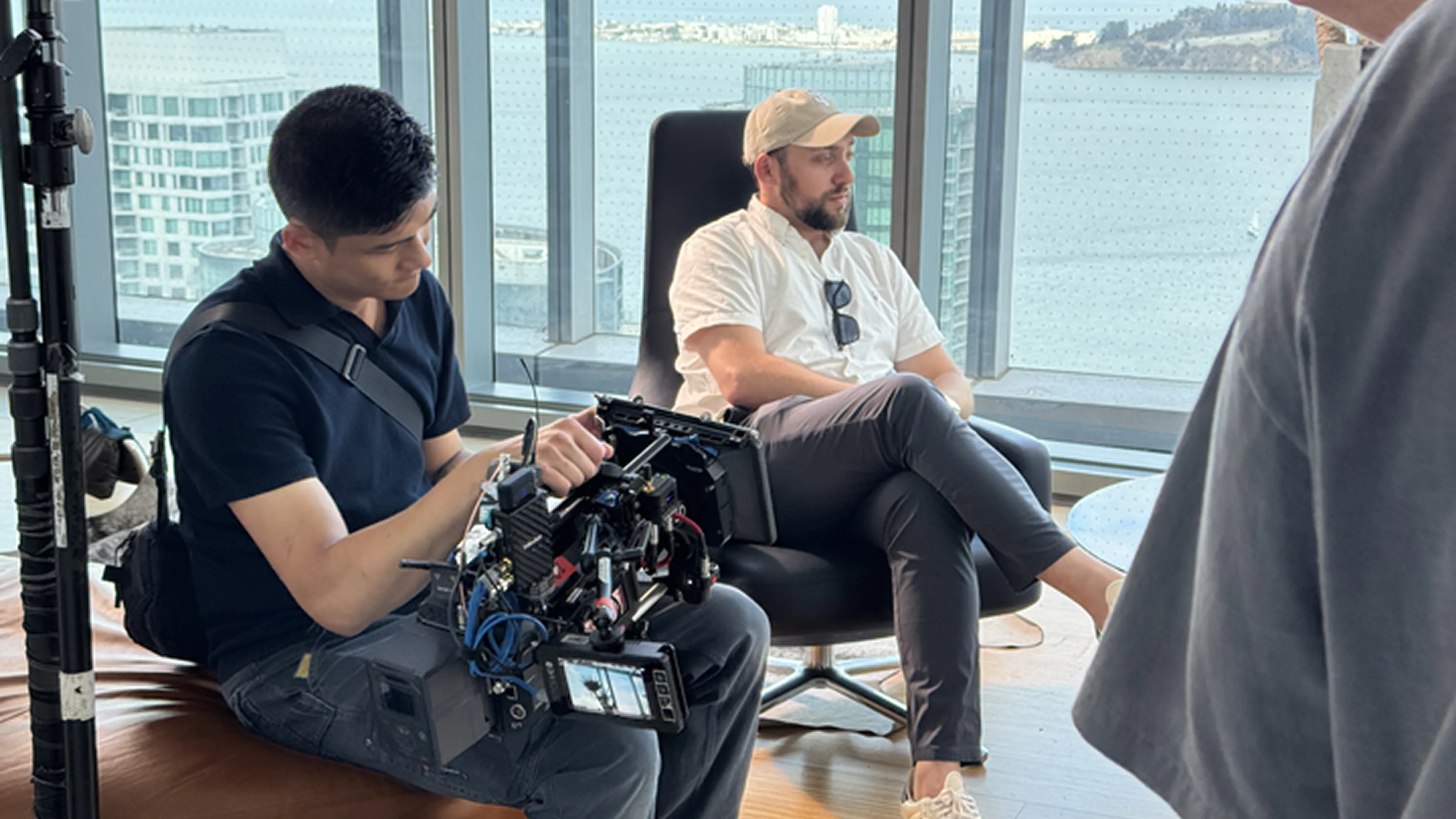 Man operating professional camera equipment indoors while another man in casual clothes sits in a chair near large windows with a waterfront view.