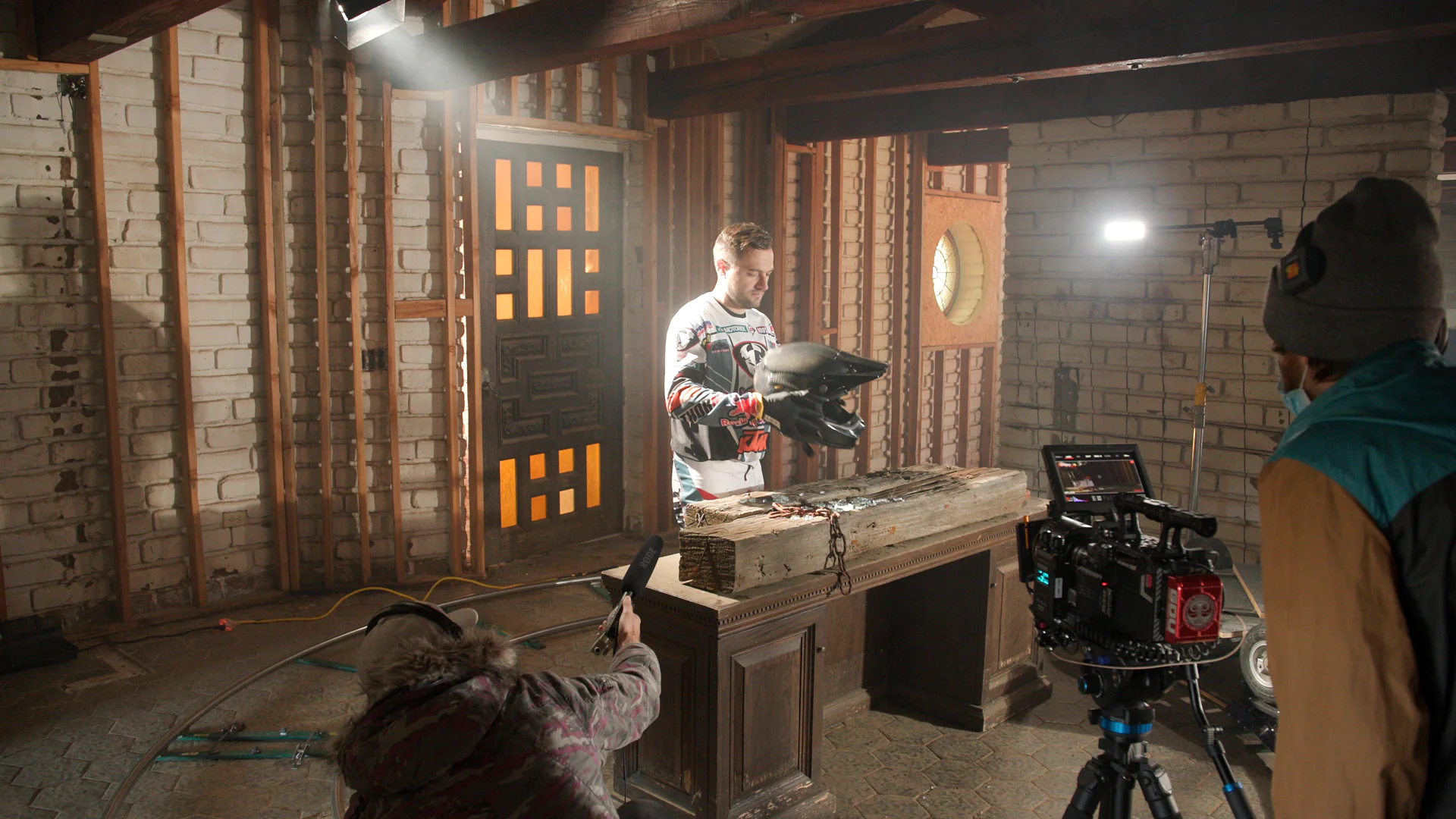 Man in motocross gear holding two black helmets standing behind a wooden desk in an unfinished room while film crew records him with professional camera and boom microphone.