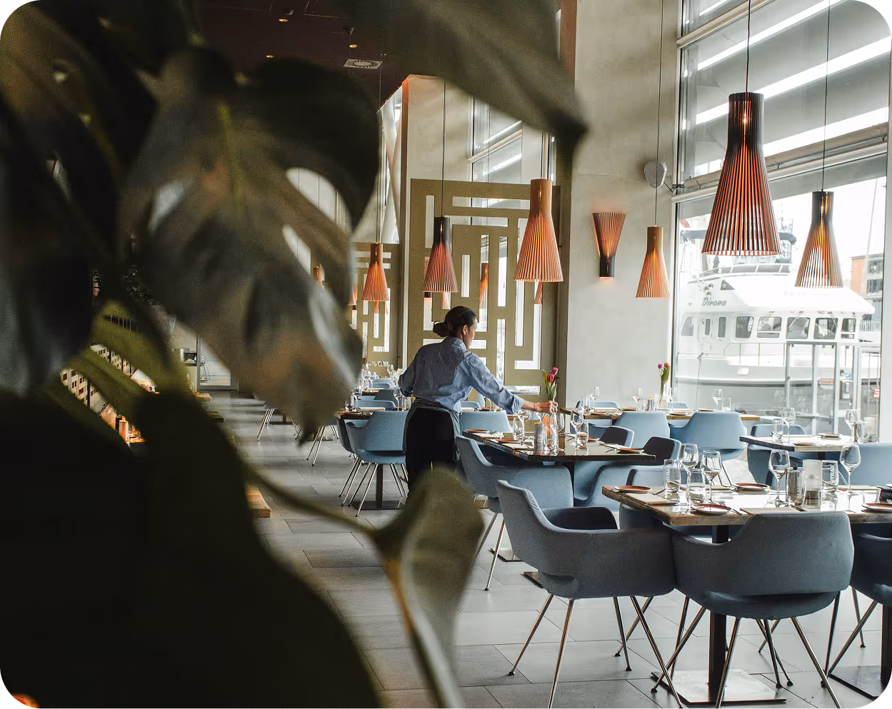 Bedienung bereitet Tische in einem modernen Restaurant mit blauen Stühlen und großen Fenstern mit Blick auf ein Boot vor.