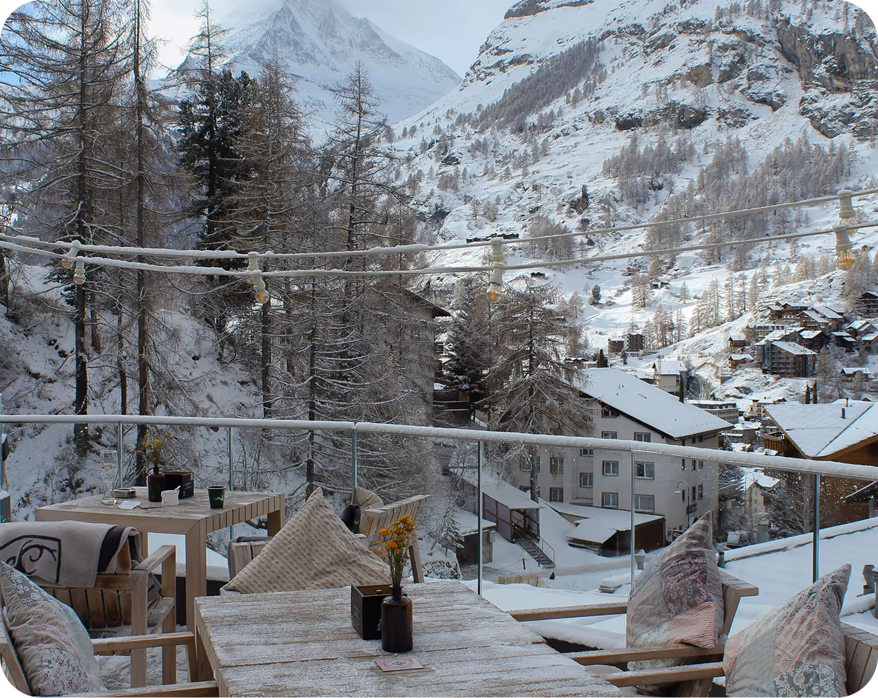 Balkon mit Holztisch, Stühlen und Kissen, schneebedeckte Bäume und Häuser vor verschneiten Bergen im Winter.
