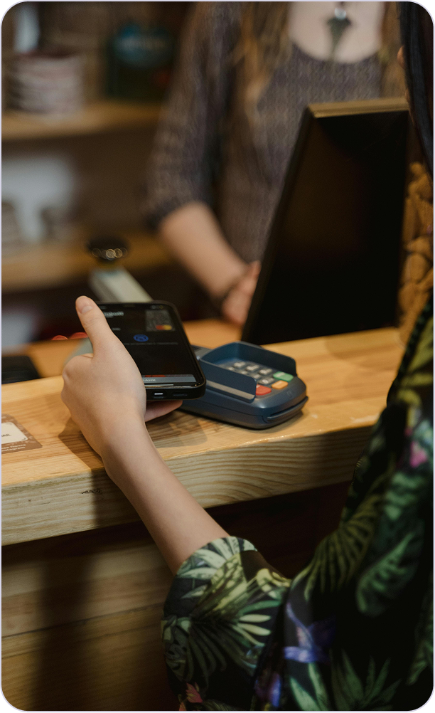 Eine Person hält ein Smartphone über ein Kartenlesegerät an einem Holztresen zur kontaktlosen Bezahlung.