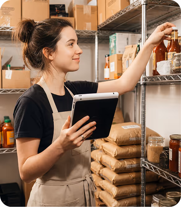 Junge Frau mit Schürze nimmt eine Flasche von einem Metallregal und hält ein Tablet in der anderen Hand in einem Lagerraum mit verschiedenen Lebensmitteln.