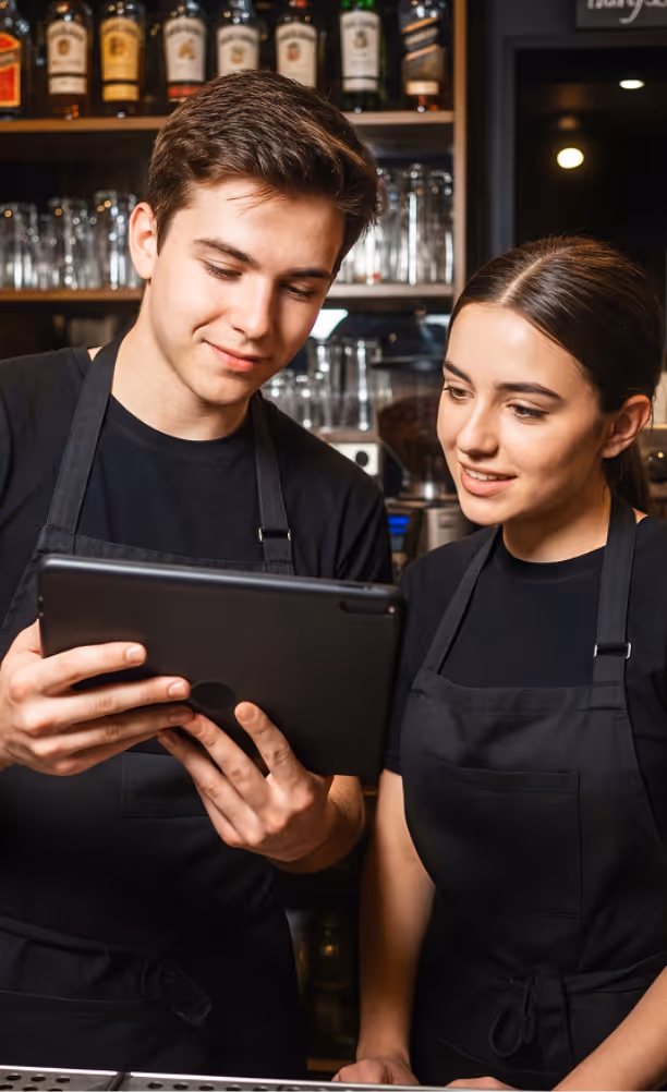 Eine Person hält ein Smartphone über ein Kartenlesegerät an einem Holztresen zur kontaktlosen Bezahlung.