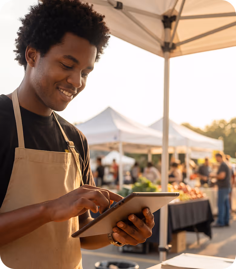 Junger Mann mit Schürze benutzt ein Tablet auf einem Freiluftmarkt mit Ständen im Hintergrund.