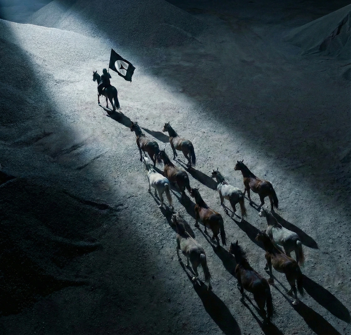 Ein Reiter hält eine comote Fahne und führt eine Gruppe von Pferden, die im Licht auf einem dunklen, felsigen Gelände laufen.