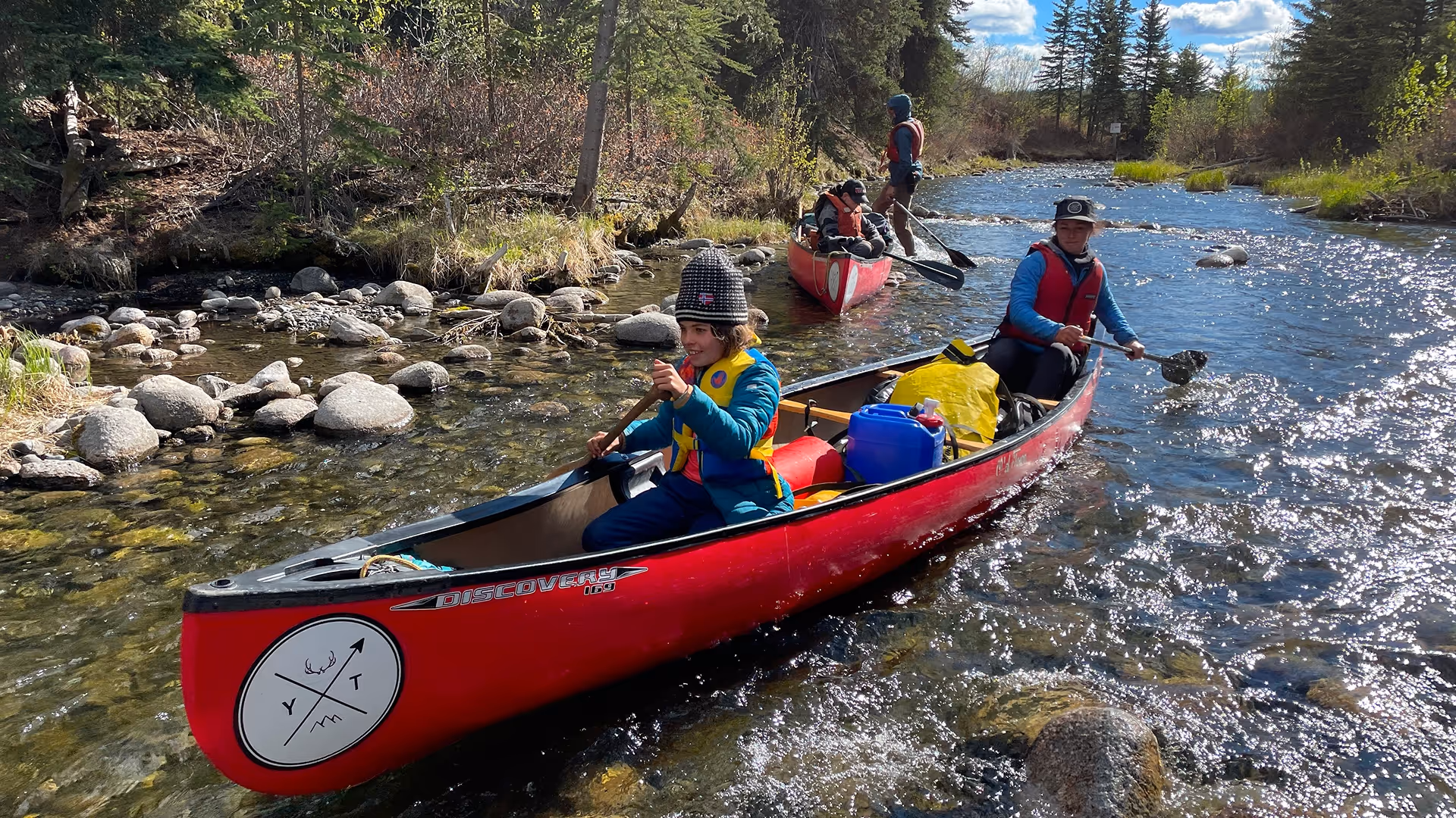 Two red canoes with WILDE School of Yukon students paddling in a shallow, rocky river near Whitehorse.
