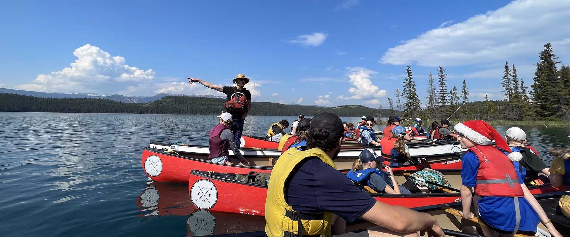 Group of WILDE School Yukon Campers in red canoes on a calm lake surrounded by forested hills under a blue sky.