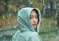 Young woman in a light blue raincoat with hood up, looking back over her shoulder while standing in the rain, with blurred nature in the background.