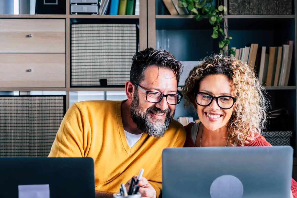 Smiling man and woman wearing glasses looking at a laptop screen together in a cozy office space.
