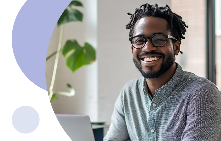 Hombre sonriente con gafas y rastas sentado en un escritorio en una habitación bien iluminada con un ordenador portátil.
