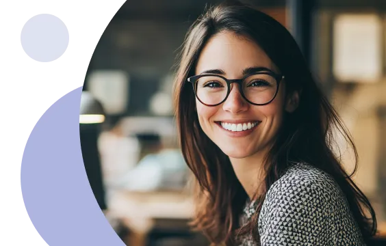 Femme souriante portant des lunettes et ayant de longs cheveux bruns, assise à l'intérieur avec un arrière-plan flou.