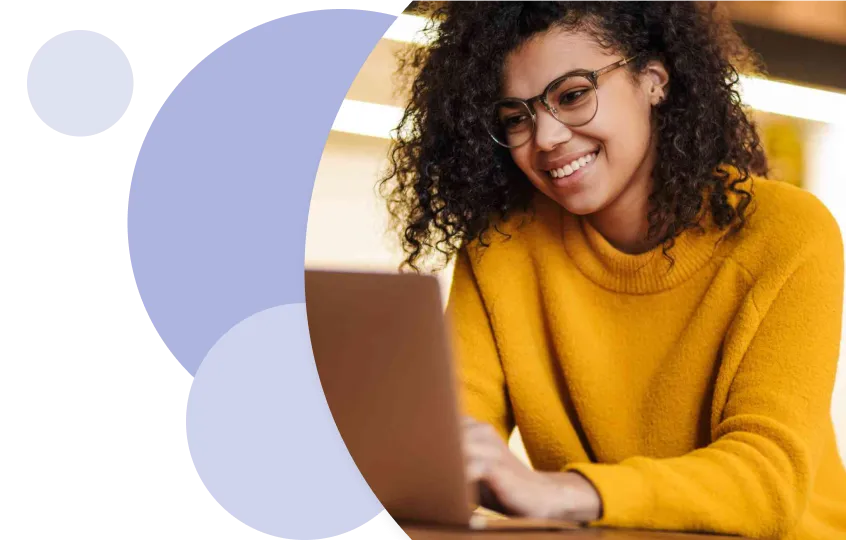 Smiling woman with curly hair and glasses working on a laptop, wearing a yellow sweater.