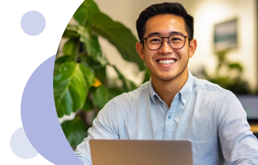 Jovem sorridente usando óculos e uma camisa azul clara sentado em uma mesa com um laptop, com grandes plantas verdes ao fundo.