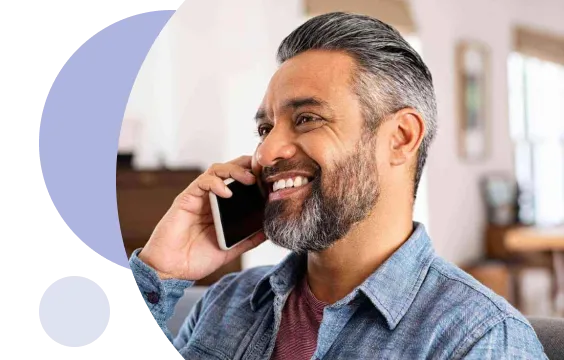 Hombre de mediana edad, con cabello y barba grises, sonriendo mientras habla por su teléfono inteligente en un interior.