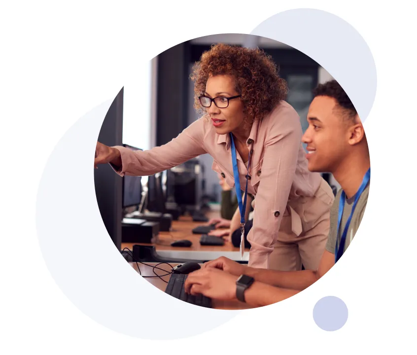 Un enseignant portant des lunettes montre un écran d'ordinateur tandis qu'un élève tape sur un clavier dans une salle informatique.