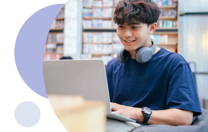 Jeune homme portant des écouteurs autour du cou travaillant sur un ordinateur portable dans une bibliothèque.