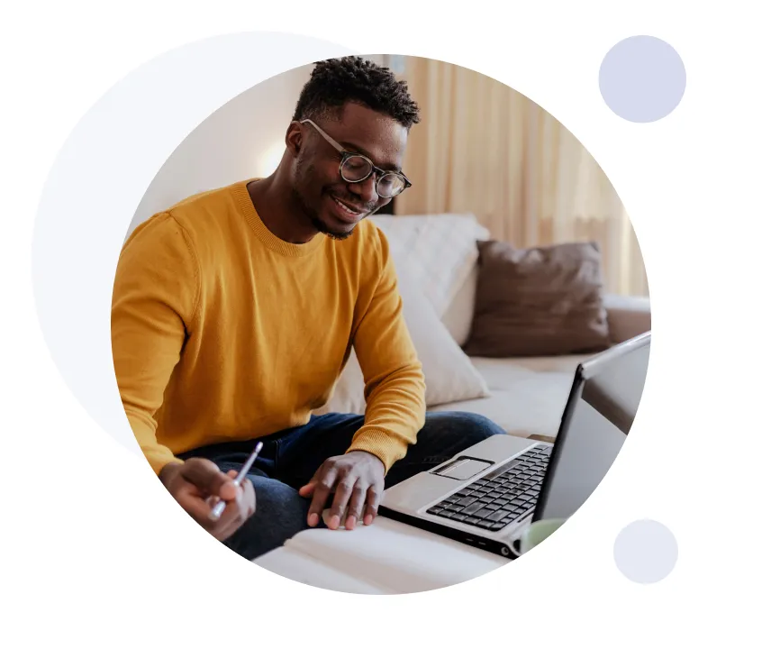 Jeune homme souriant portant des lunettes et un pull moutarde, utilisant un ordinateur portable et écrivant dans un cahier sur un canapé.
