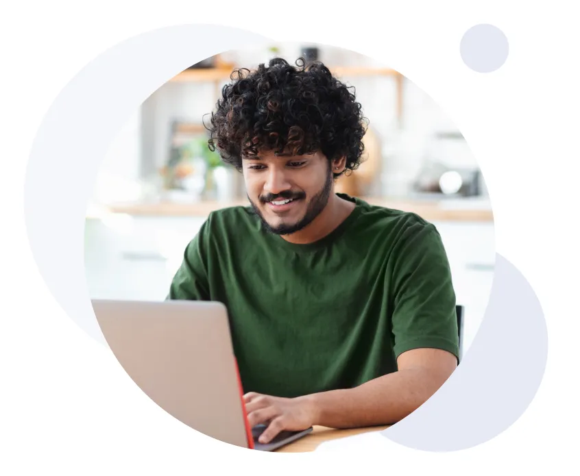 Jeune homme souriant aux cheveux bouclés travaillant sur un ordinateur portable à l'intérieur.