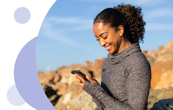 Mujer sonriente con cabello rizado y camiseta gris de manga larga mirando su smartphone al aire libre, con rocas y un cielo azul al fondo.