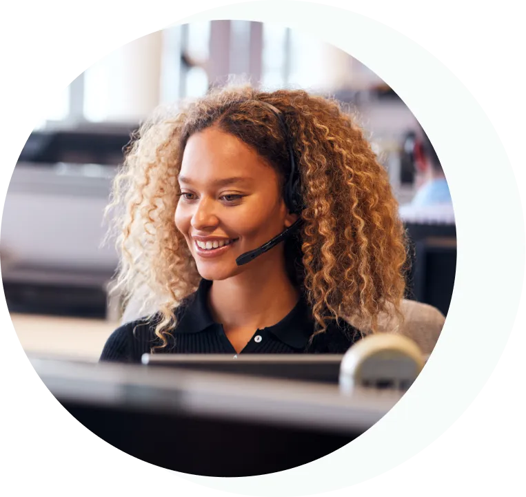 Femme souriante aux cheveux bouclés portant un casque audio travaillant sur un ordinateur dans un bureau.