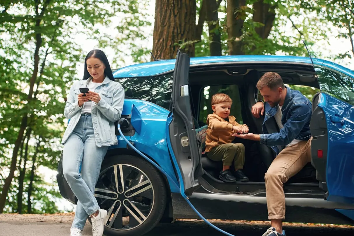 Une femme appuyée contre une voiture électrique bleue pendant qu'elle se recharge, un homme et un enfant assis à l'intérieur de la voiture, la porte ouverte.