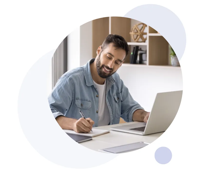 Homme barbu portant une chemise en jean, souriant tout en écrivant dans un cahier, assis à une table avec un ordinateur portable ouvert dans un bureau moderne à domicile.