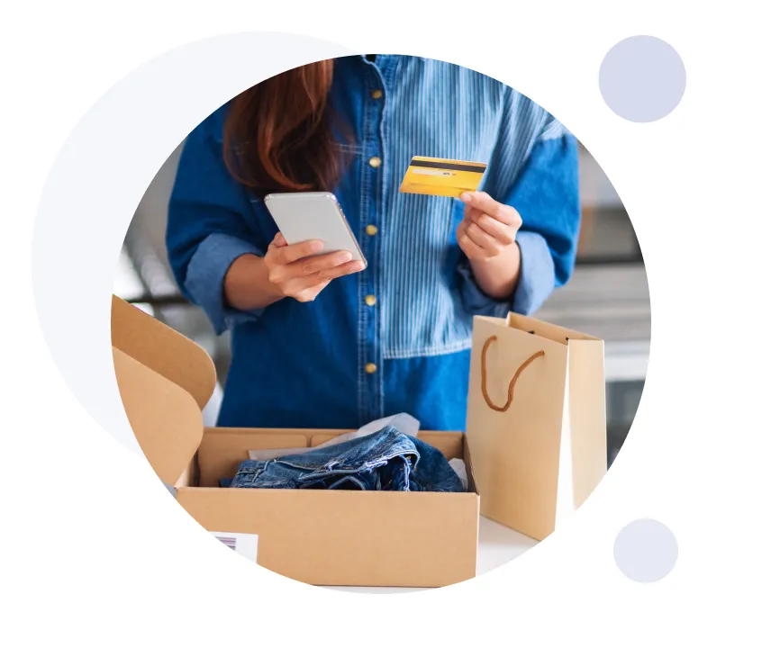 Person holding a smartphone and a credit card near an open box with folded denim jeans and a paper shopping bag on the table.