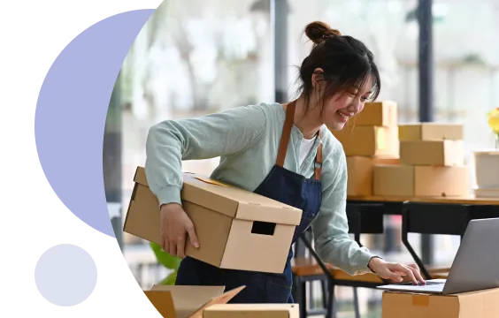 Woman in apron holding a cardboard box and working on a laptop surrounded by stacked boxes in a bright workspace.