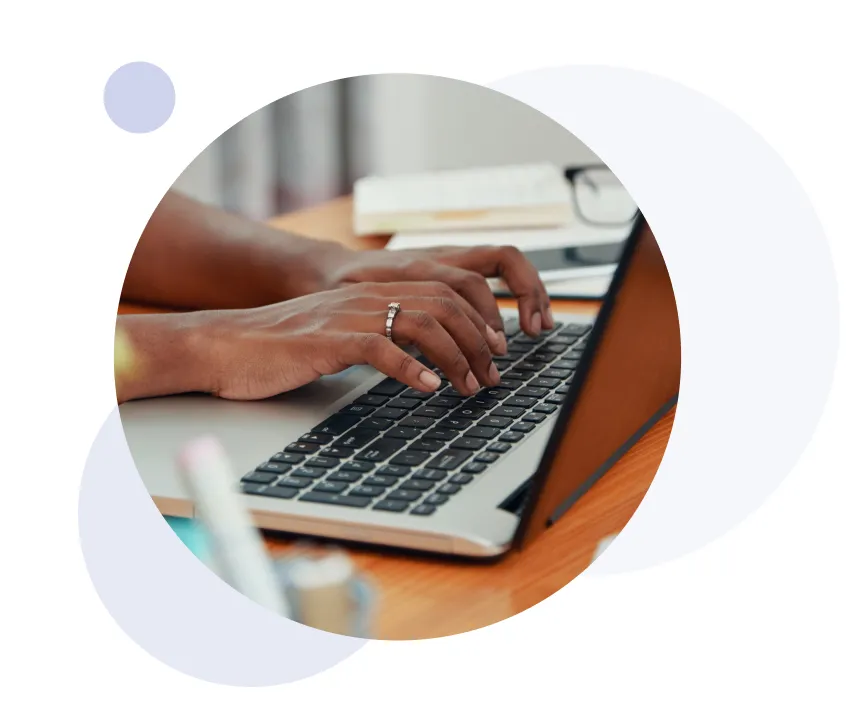 Close-up of hands typing on a laptop keyboard with a ring on one finger, placed on a wooden desk.