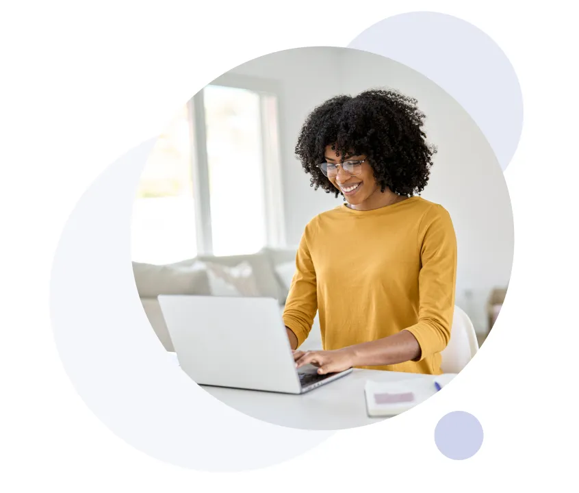 Smiling woman with curly hair and glasses typing on a laptop at a white desk in a bright room.