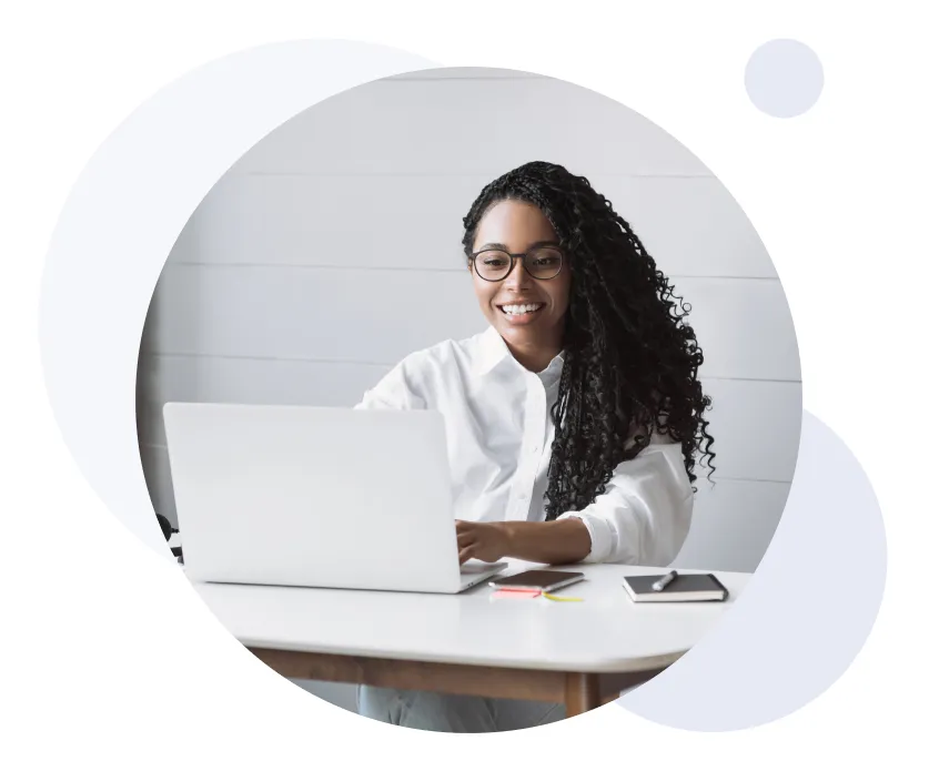Mujer sonriente con cabello rizado y gafas trabajando en un ordenador portátil en un escritorio blanco con un cuaderno y un teléfono.