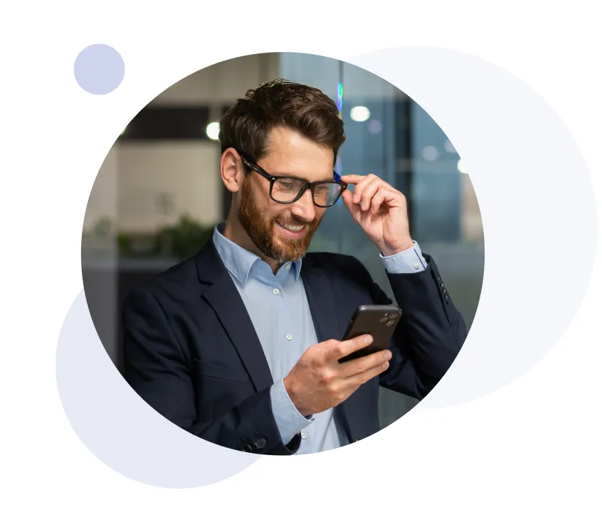 Hombre barbudo sonriente con gafas y traje mirando su smartphone en interiores.