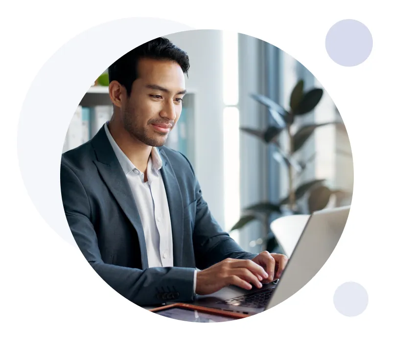 Man in business attire typing on a laptop in a modern office setting with large windows and plants.