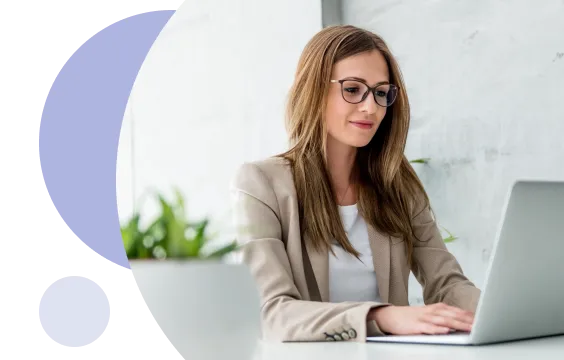 Young woman wearing glasses and beige blazer working on a laptop at a white desk with a blurred plant in the background.