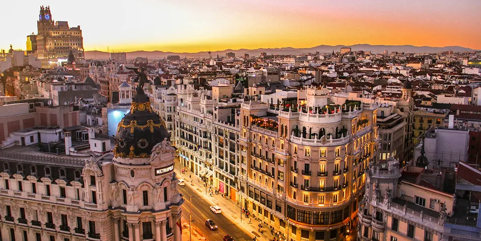 Vista noturna da paisagem urbana de Madri com o edifício Metropolis e ruas iluminadas sob um céu dourado ao pôr do sol.