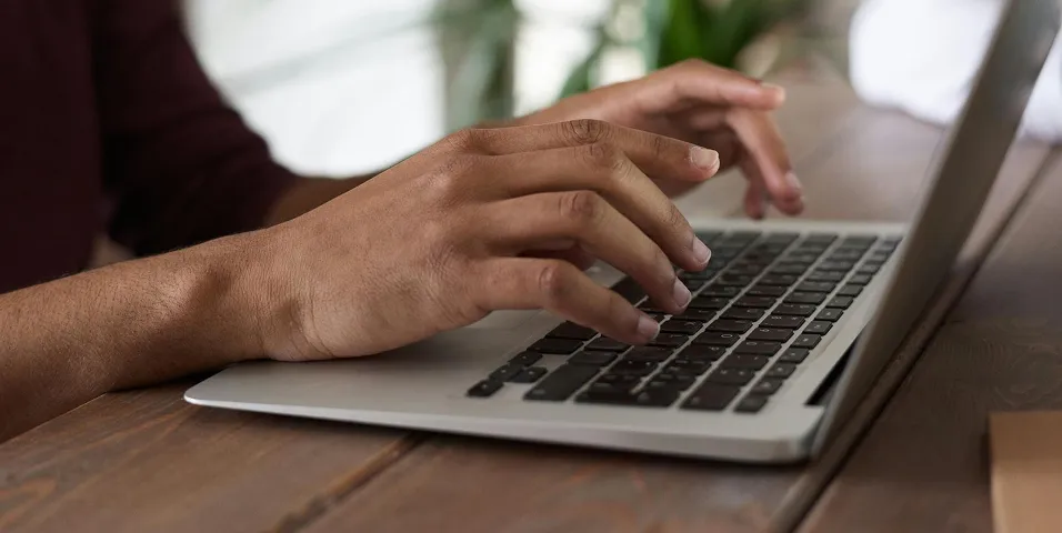 Close-up de mãos digitando no teclado de um laptop sobre uma mesa de madeira.